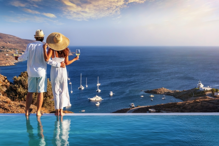 Ein sich umarmendes, verliebtes Paar genießt im Urlaub den Sommer-Sonnenuntergang über der Ägäis am Swimmingpool bei einem Drink.